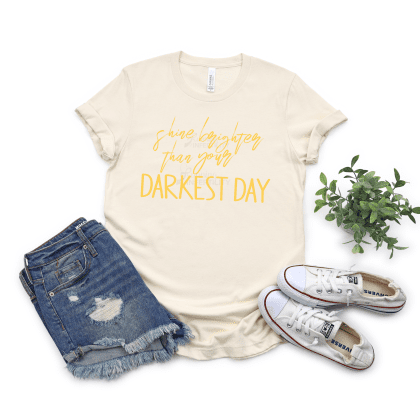 Shine Brighter than your darkest Day cursive and straight yellow letters on a heather natural tee