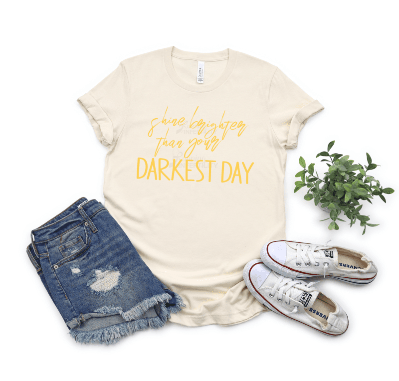 Shine Brighter than your darkest Day cursive and straight yellow letters on a heather natural tee
